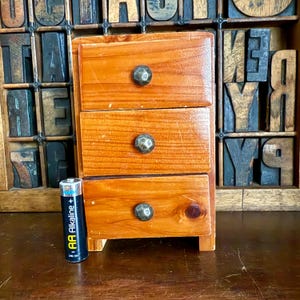 May include: A small, three-drawer wooden cabinet with round, dark metal knobs. The cabinet is made of light brown wood and has a rustic appearance. An AA battery is placed next to the cabinet for size comparison.