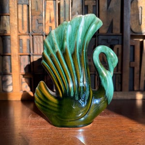 May include: A ceramic swan-shaped vase with a glossy, multi-toned glaze. The vase features a fan-shaped back and a curved neck, with shades of green and brown. The vintage decorative piece is likely for home decor.