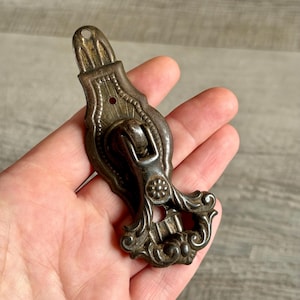 Könnte beinhalten: Ein antiker, verzierter Metall-Schubladengriff mit einem dekorativen Blumendesign. Der Griff hat eine silberfarbene Patina mit filigranen Details und einer kleinen Rolle. Die Oberseite hat ein Loch zur Montage.