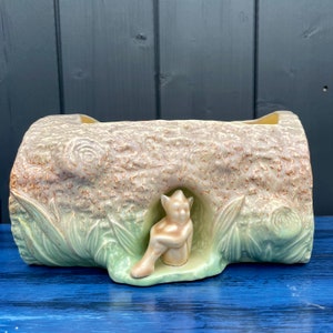May include: A ceramic planter shaped like a tree trunk with a small fairy figurine sitting inside. The planter is a light green and brown colour with a textured surface.