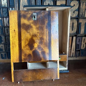 May include: A vintage wooden cabinet with a hinged door and a small drawer. The cabinet is made of dark wood with a natural grain pattern. The door is slightly ajar, revealing the interior. The cabinet is likely used for storage.