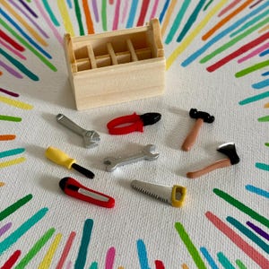 Könnte beinhalten: Eine Holzkiste mit verschiedenen Miniaturwerkzeugen: Schraubenschlüssel, Zange, Schraubendreher, Hammer, Axt, Säge und Cutter. Die Werkzeuge sind silber, rot, gelb und braun. Der Hintergrund ist weiß mit bunten diagonalen Linien.