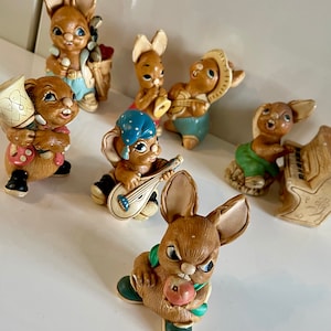 May include: A collection of seven ceramic figurines of bunnies playing musical instruments. The bunnies are painted in various colors, including brown, white, blue, green, and yellow. The figurines are all wearing different outfits and have different expressions on their faces.
