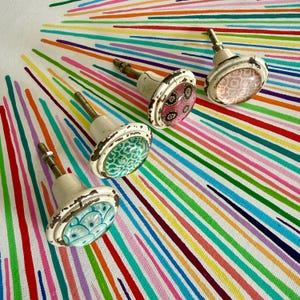 May include: Four round cabinet knobs with distressed white metal bases and threaded metal stems. Each knob features a unique circular inlay with distinct patterns: a light blue fan design, a green floral print, a pink floral motif, and a light pink geometric pattern.
