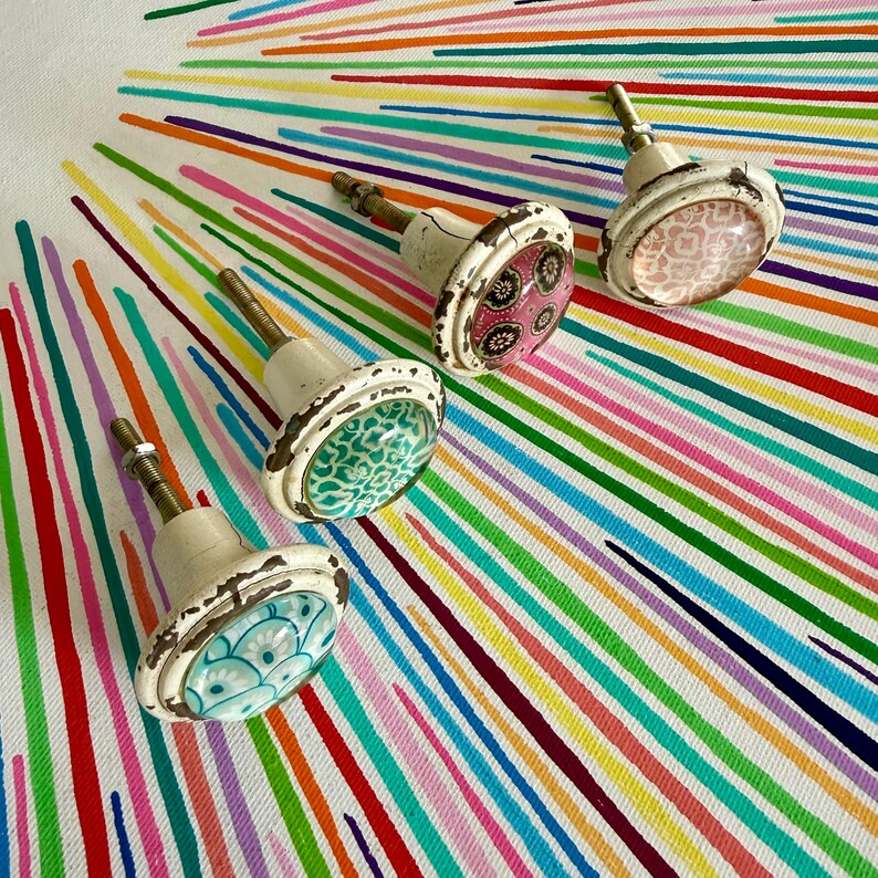 May include: Four round cabinet knobs with distressed white metal bases and threaded metal stems. Each knob features a unique circular inlay with distinct patterns: a light blue fan design, a green floral print, a pink floral motif, and a light pink geometric pattern.