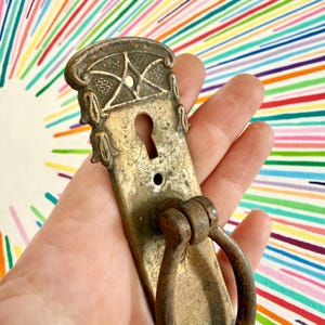 May include: An antique brass door escutcheon plate featuring a weathered patina, a central keyhole, and a circular drop handle. The top section displays an intricate embossed geometric pattern with a small screw hole for mounting purposes.