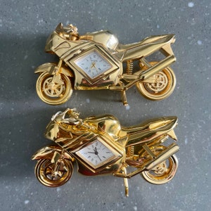 Könnte beinhalten: Zwei goldfarbene Miniatur-Motorrad-Uhren. Jedes Motorrad hat ein rundes Zifferblatt mit schwarzen Zeigern und römischen Ziffern. Die Motorräder sind mit einer Fahrerfigur und einem schwarzen Sitz detailliert.