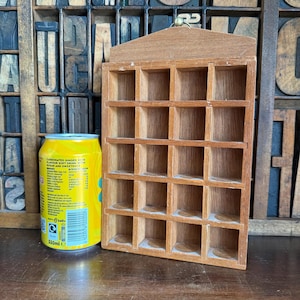 May include: A rectangular wooden display shelf featuring twenty individual square compartments arranged in a four-by-five grid, topped with a triangular peak and a small metal hanging loop, positioned next to a yellow aluminium soda can.