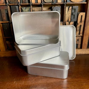 May include: Four silver metal rectangular boxes stacked on top of each other. The boxes are empty and have a smooth, shiny surface.