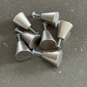 May include: Seven brushed nickel cabinet knobs are displayed. Each knob features a conical design with a flat, circular base and a screw for attachment. The knobs are arranged on a grey surface.