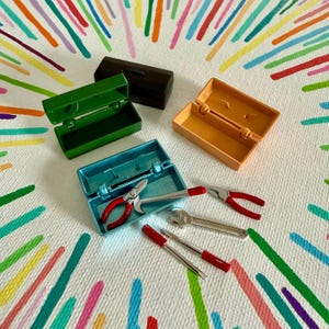 Könnte beinhalten: Miniatur-Werkzeugkästen in Grün, Braun, Blau und Orange sind geöffnet und zeigen kleine Werkzeuge. Zu den Werkzeugen gehören Zangen, ein Schraubenschlüssel und Schraubendreher. Der Hintergrund ist eine weiße Oberfläche mit bunten, gemalten Linien.