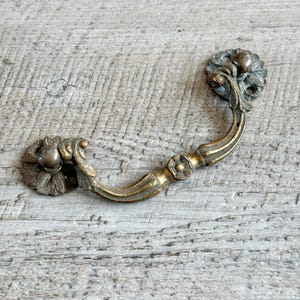 Könnte beinhalten: Ein antiker Schubladengriff aus Messing mit aufwendigen floralen Details. Der Griff hat einen gebogenen Griff und zwei dekorative Rosetten. Das Messing hat ein verwittertes Aussehen, was ihm einen Vintage-Charme verleiht. Ideal, um Möbeln einen Hauch von Eleganz zu verleihen.