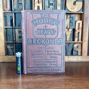 May include: A brown leather-bound book titled "The Model Ready Reckoner" with tables of interest, bank rate, commission, stamp duties, exchange, wages, weights & measures, commercial forms, and 40,000 calculations.