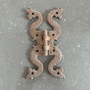 Könnte beinhalten: Ein dekoratives Scharnier im antiken Stil aus verwittertem Metall. Das Scharnier hat ein symmetrisches Design mit geschwungenen, scrollartigen Formen und dekorativen Aussparungen. Das Metall hat ein rostiges, gealtertes Aussehen, was darauf hindeutet, dass es Vintage ist oder den Elementen ausgesetzt war.