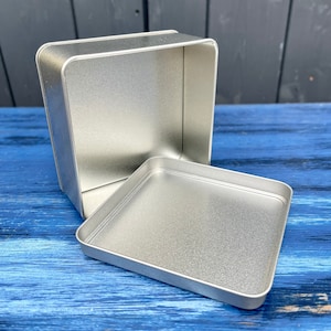 May include: A silver metal square tin box with the lid open. The tin is empty and has a smooth, shiny surface.