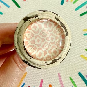 May include: A round cabinet knob featuring a distressed off-white metal frame with visible cracks and dark edges. The center is inlaid with a smooth, glossy glass dome displaying a repeating pale pink and white geometric quatrefoil pattern.