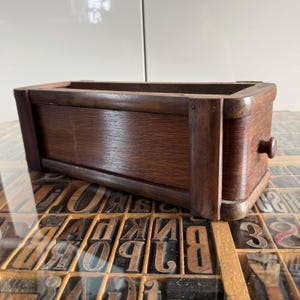 May include: A rectangular wooden box with a dark brown finish, featuring a smooth front panel, reinforced vertical corner supports, and a small, centred circular knob on the right side. The box has an open top and a sturdy, structured frame.