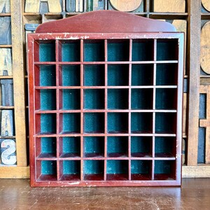 May include: A square wooden display case with a dark cherry finish, featuring a grid of thirty-six small square compartments. The back of each individual cubby is lined with dark green felt, and the frame shows minor signs of wear on the edges.