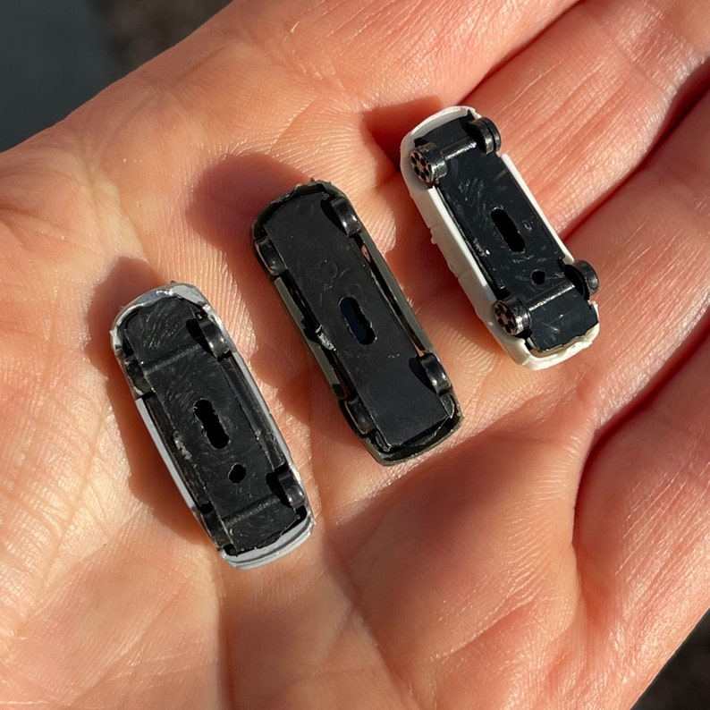 May include: Three miniature toy cars, two white and one black, with black wheels and detailed features. The cars are arranged on a hand.