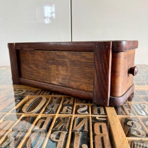 Peut inclure: Une boîte de rangement rectangulaire en bois avec une finition brun foncé et un petit bouton sur le côté. La boîte présente un grain de bois visible et est construite avec des supports d'angle verticaux. Elle repose sur une surface plane.
