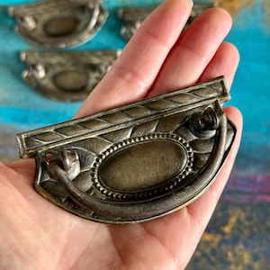 May include: Antique bronze-toned drawer pulls with a decorative design. The pulls feature an oval center and a curved handle. The background shows additional pulls, suggesting a set. The design includes a rope-like border and a beaded oval center.