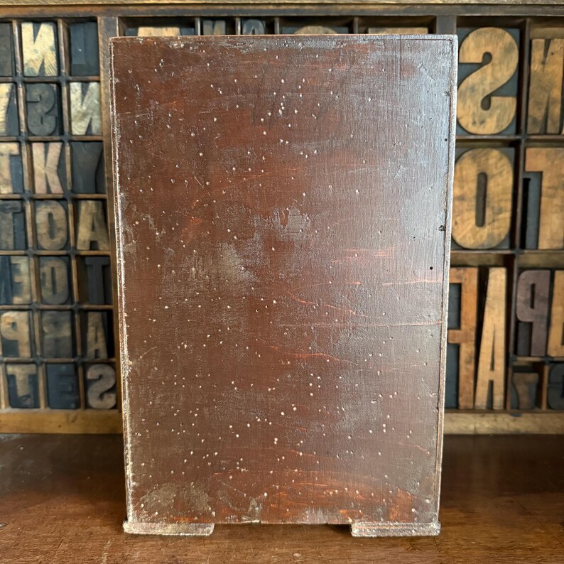 May include: A rectangular, wooden box with a dark brown finish and visible wood grain. The box has a slightly distressed appearance with small, white specks scattered across its surface. The background features vintage wooden letterpress type blocks.
