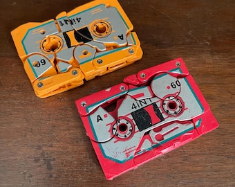 May include: Two vintage cassette tapes, one yellow and one red, with the text "4INT" and "60" on them. The tapes are in a folded, compact design.