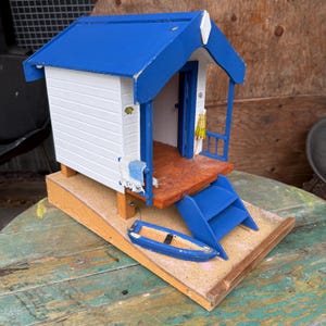 May include: A detailed miniature beach hut model, painted white with blue accents and a blue roof. Features include a small blue boat, a wooden deck, and blue stairs. The hut's door is open, offering a view of the interior.