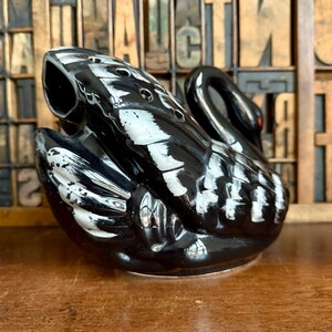 May include: A black ceramic swan-shaped decorative object with white accents. The swan has a long neck and a curved body, with a small red dot for an eye. The object is sitting on a wooden surface.