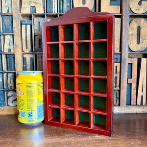 May include: A rectangular wooden display case with a dark reddish-brown finish and a green felt-lined back. The case features a grid of twenty-four small square compartments arranged in four columns and six rows, positioned next to a 330ml soda can.