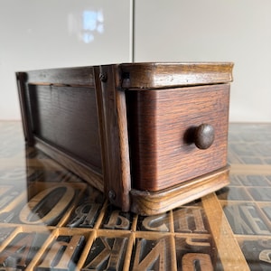 May include: A rectangular dark brown wooden storage box featuring a single sliding drawer with a small round wooden knob. The box is constructed with visible wood grain and secured with metal screws on the side frame, showcasing a smooth finish.