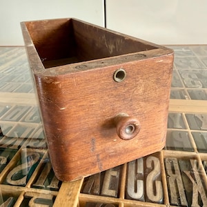 Könnte beinhalten: Eine rechteckige Holzschublade mit mittelbraunem Finish, ausgestattet mit einem einzelnen runden Holzknauf an der Vorderseite und einer kleinen kreisförmigen Metallöse nahe der Oberkante. Die Schublade zeigt sichtbare Maserung und Abnutzung.