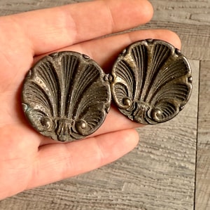May include: Two antique, round, metal shell-shaped buttons with a detailed fan design. The buttons are a tarnished bronze colour, showing signs of age and wear. The buttons are held in a hand.