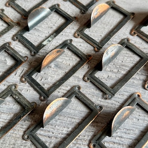 Könnte beinhalten: Nahaufnahme mehrerer Vintage-Metall-Etikettenhalter. Jeder Halter hat einen rechteckigen Rahmen mit abgerundeten Ecken und einen gebogenen Etikettenhalter. Das Metall hat ein abgenutztes, gealtertes Aussehen mit Rostansätzen.