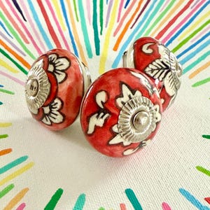 May include: Three red ceramic cabinet knobs with white floral designs and silver-coloured hardware. The knobs are oval-shaped and have a glossy finish. The background is a colourful, abstract pattern.