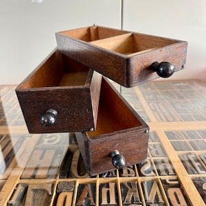 May include: Three antique wooden drawers with dark brown exteriors and black knobs. The drawers are stacked at an angle, revealing their empty interiors. The drawers are resting on a surface with letterpress printing blocks.
