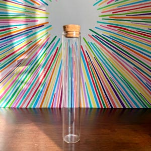 May include: A clear glass vial with a cork stopper. The cylindrical vial is displayed against a backdrop of radiating lines in a variety of colours. The background is a burst of rainbow hues.
