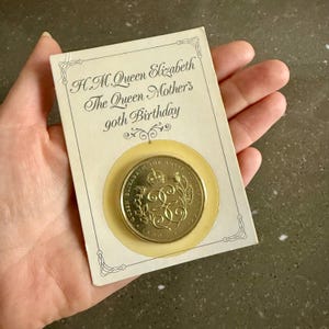 May include: A gold-coloured commemorative coin for H.M. Queen Elizabeth, The Queen Mother's 90th Birthday, set on a cream card. The card has decorative borders and text that reads "H.M. Queen Elizabeth The Queen Mother's 90th Birthday."