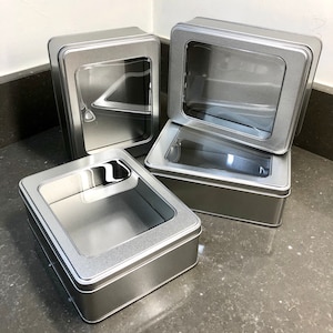May include: Four rectangular silver metal tins with clear plastic windows. One tin has its lid open, revealing the interior. The tins are arranged on a grey surface, showcasing their design and potential for storage or display.