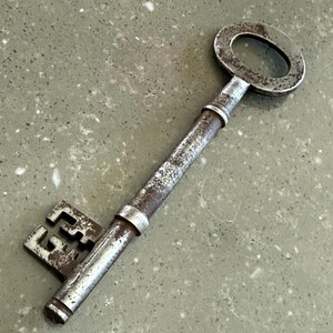 Könnte beinhalten: Ein antiker Metallschlüssel mit einem großen, ovalen Griff und einem detaillierten Bart. Der Schlüssel weist Alterserscheinungen mit einem rostigen, verwitterten Aussehen auf. Der Schaft des Schlüssels ist zylindrisch, und der Bart hat ein komplexes, geometrisches Design.