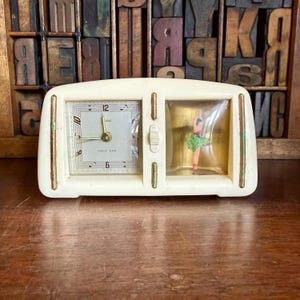 May include: Cream-coloured vintage clock with a square clock face and a separate compartment with a ballerina figurine. The clock face has black numerals and hands. The ballerina is wearing a green tutu. The clock has the words "Music Alarm".
