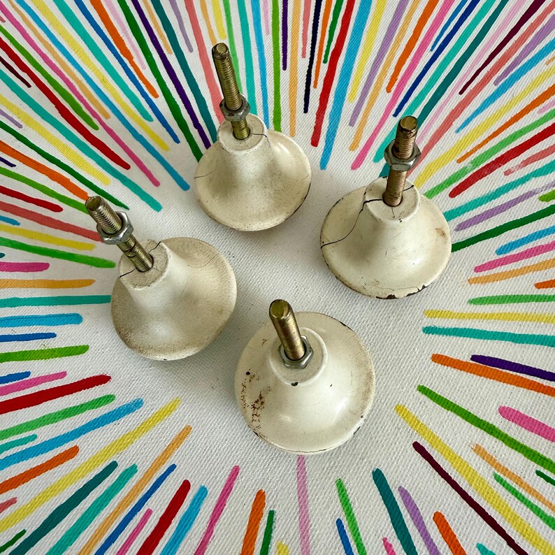 May include: Four off-white, bell-shaped cabinet knobs with visible surface cracks and wear. Each knob features a central threaded metal bolt extending upward, secured by a small hexagonal metal nut. The knobs have a smooth, rounded base.