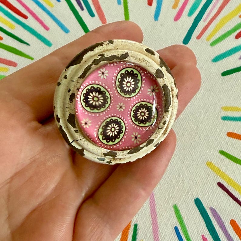 May include: A circular knob featuring a distressed, cream-colored outer rim with visible dark brown paint chips. The center is a smooth, glossy inlay displaying a vibrant pink background patterned with repeating dark brown and light green floral motifs.