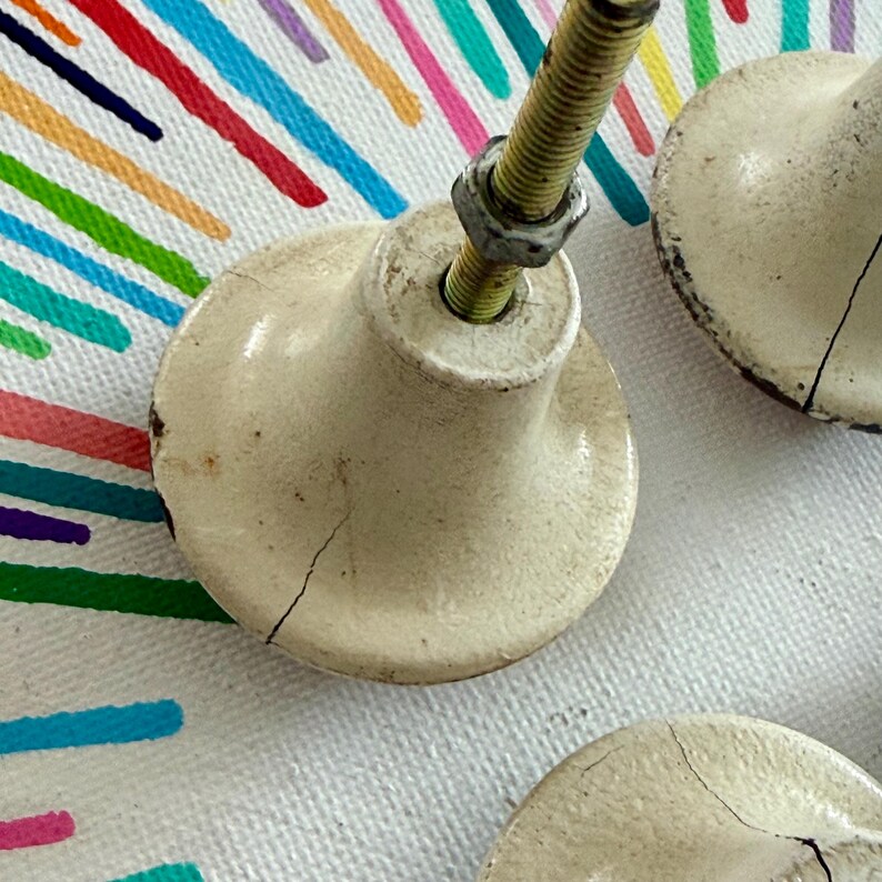 May include: Off-white ceramic cabinet knobs with a rounded, flared shape and visible surface cracks. Each knob features a central threaded metal bolt with a small hexagonal nut attached, showing signs of wear and aging on the finish.