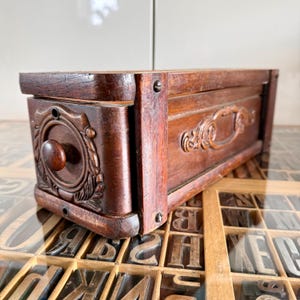 May include: A small, antique wooden box with a dark brown finish. The box features ornate carvings, including a circular design with a knob and a decorative scroll pattern. The box is rectangular and appears to be a storage container.