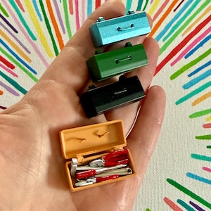 May include: Miniature toy toolboxes in teal, green, and black, with an open orange toolbox revealing tiny tools. The tools include a hammer, pliers, and a wrench. The toolboxes are held in a hand.