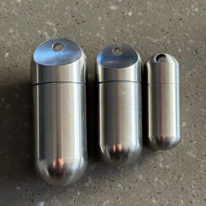 May include: Three brushed metal capsule-shaped containers of varying sizes. Each has a rounded bottom and a flat top with a small hole. The containers are silver and appear to be made of stainless steel.