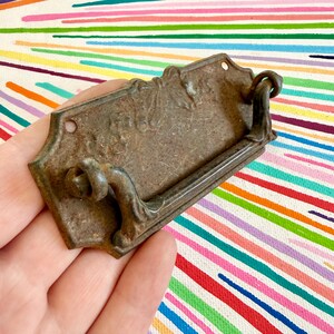 May include: A rectangular metal drawer pull with a weathered, dark brown patina and a textured surface. The hardware features a central swinging handle attached to a decorative backplate with two visible screw holes for mounting purposes.