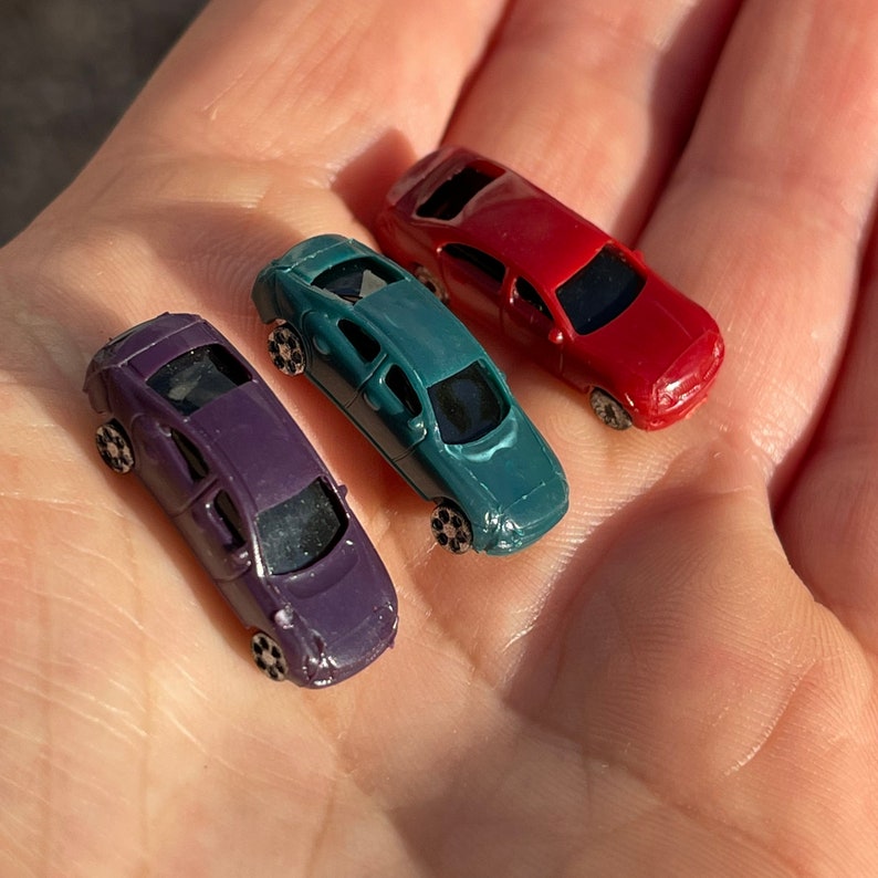 May include: Three miniature toy cars in different colors: purple, teal, and red. The cars are sitting on a person's hand.