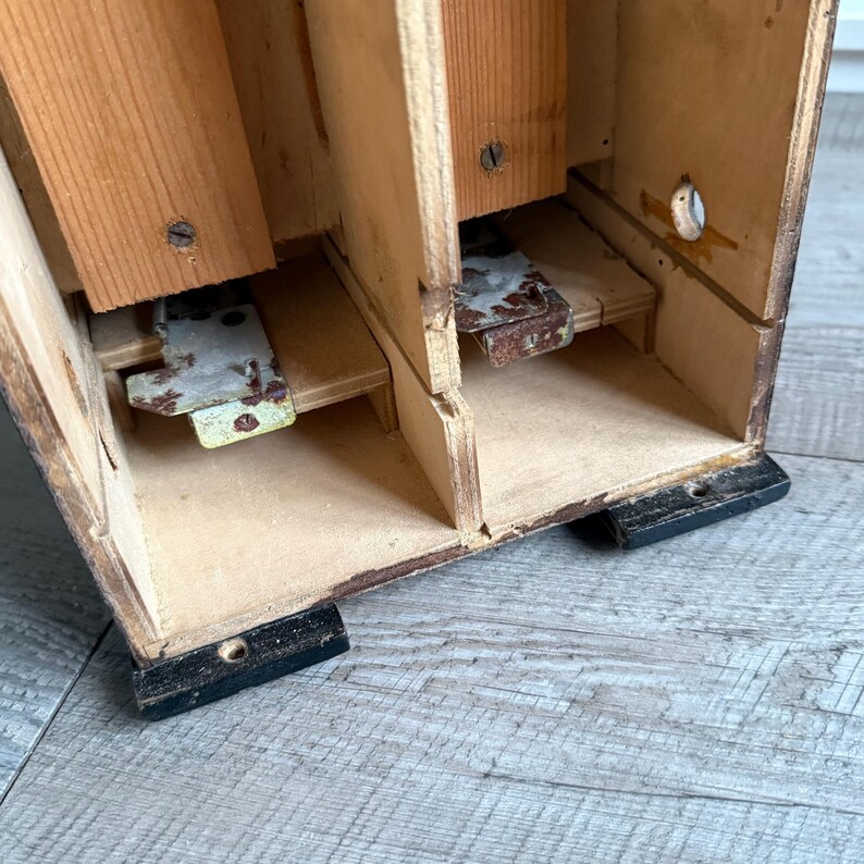 May include: A vintage wooden box with multiple compartments. The box is constructed from light-colored wood with visible grain and features metal hinges and supports. The base of the box is painted black, and the interior shows signs of age and wear.